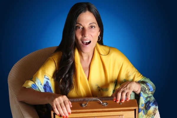 danielle blackman cartau, femme, cheveux long, brun, en train de chanter, elle joue à un instrument de musique, shrutibox, fond bleu, elle port un haut jaune et bleu