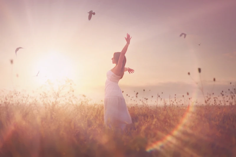 femme dans un champs les bras ouverts avec le soleil qui brille et les oiseaux qui volent autour d'elle